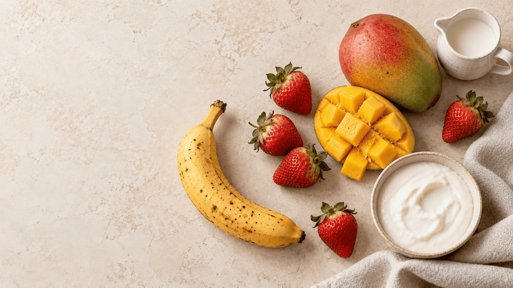 AI-created overhead ingredient illustration of a ripe banana, strawberries, whole and cut mango, plain yogurt and a milk pitcher on a cream surface with linen, leaving open space at the left.